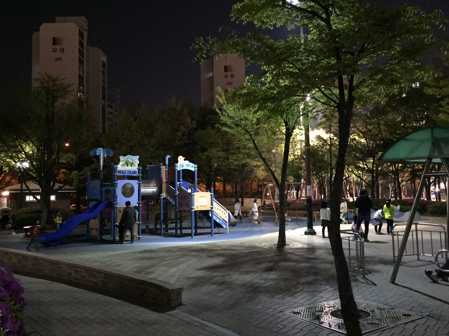 밤이 된 놀이터예요. 가로등 불빛이 놀이 기구를 비추고 아이들 여럿이 놀고 있어요. 뒤로는 조명을 받은 아파트 두 동이 서 있어요.