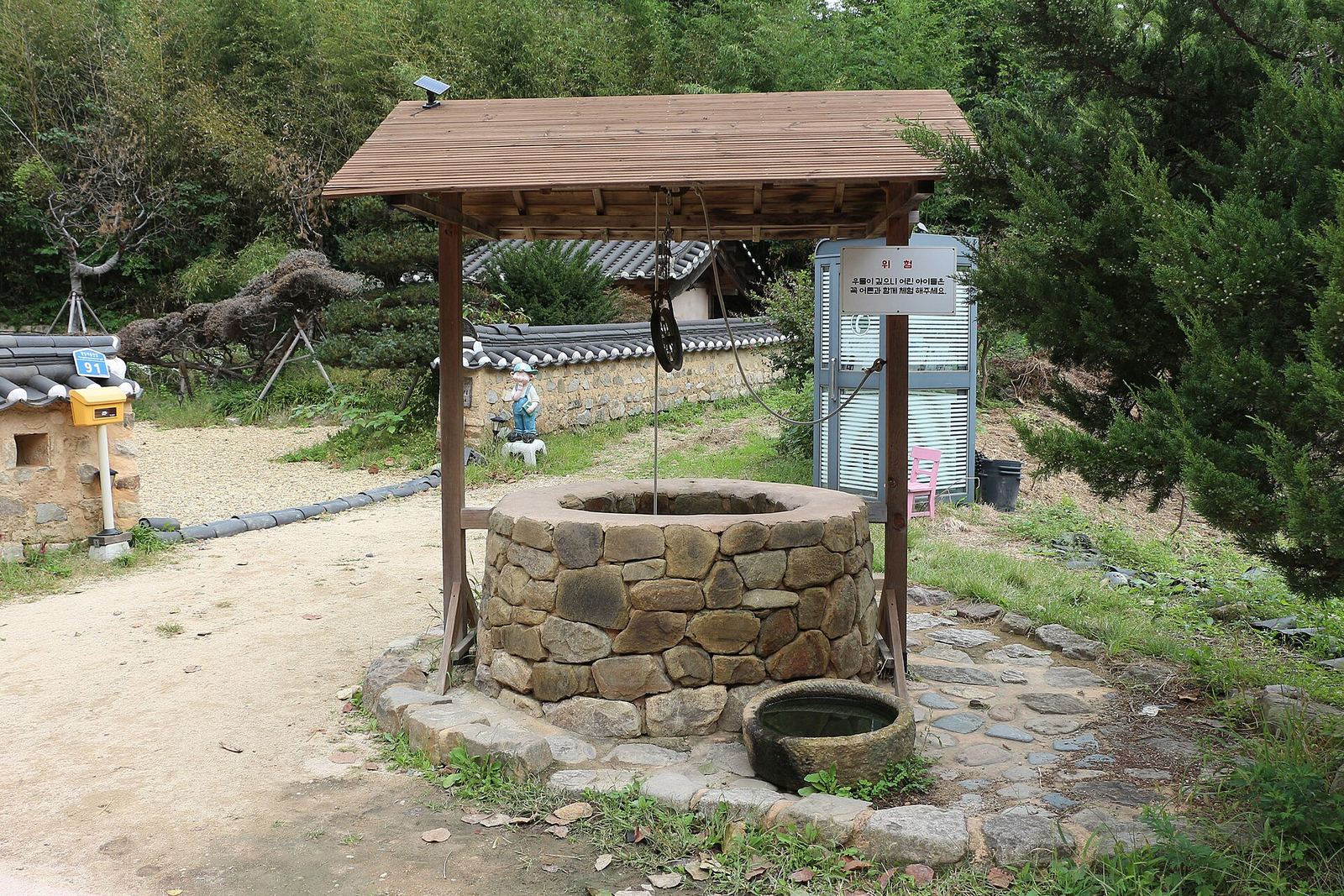 돌을 둥글게 쌓아 만든 우물이에요. 나무 지붕이 덮여 있고 줄에 매달린 도르래가 보여요.