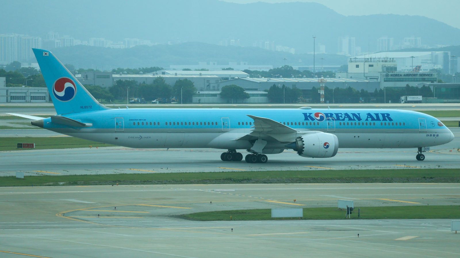 온몸이 하늘색인 큰 비행기가 바퀴를 내리고 공항 길 위에 서 있다. 뒤로 낮은 공항 건물이 늘어서 있다.