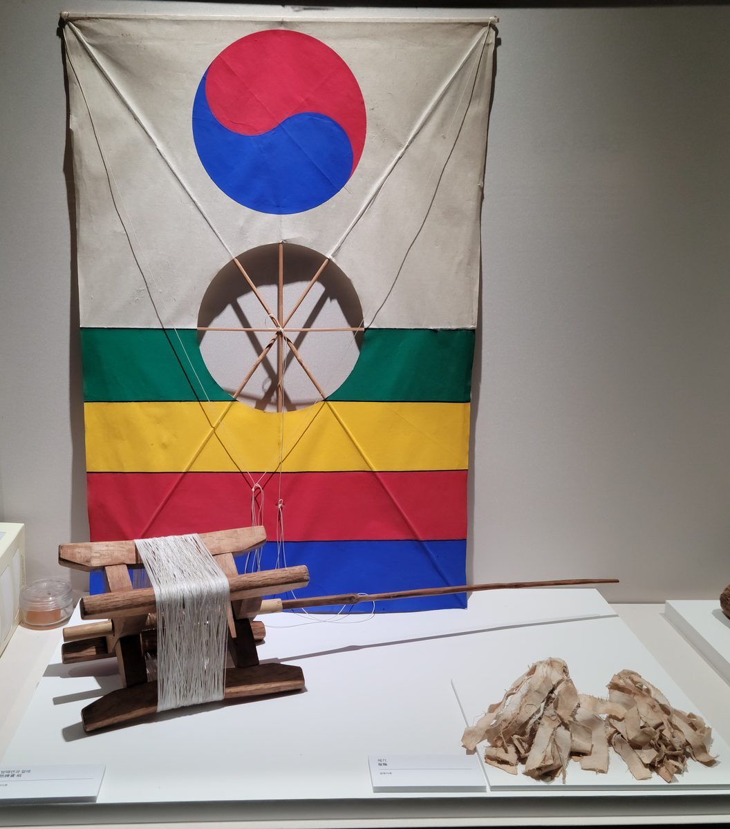 가운데에 구멍이 뚫리고 태극무늬와 초록·노랑·빨강·파랑 띠가 그려진 연이 걸려 있고, 그 앞에 실을 감은 나무 얼레와 천 제기가 놓여 있다.