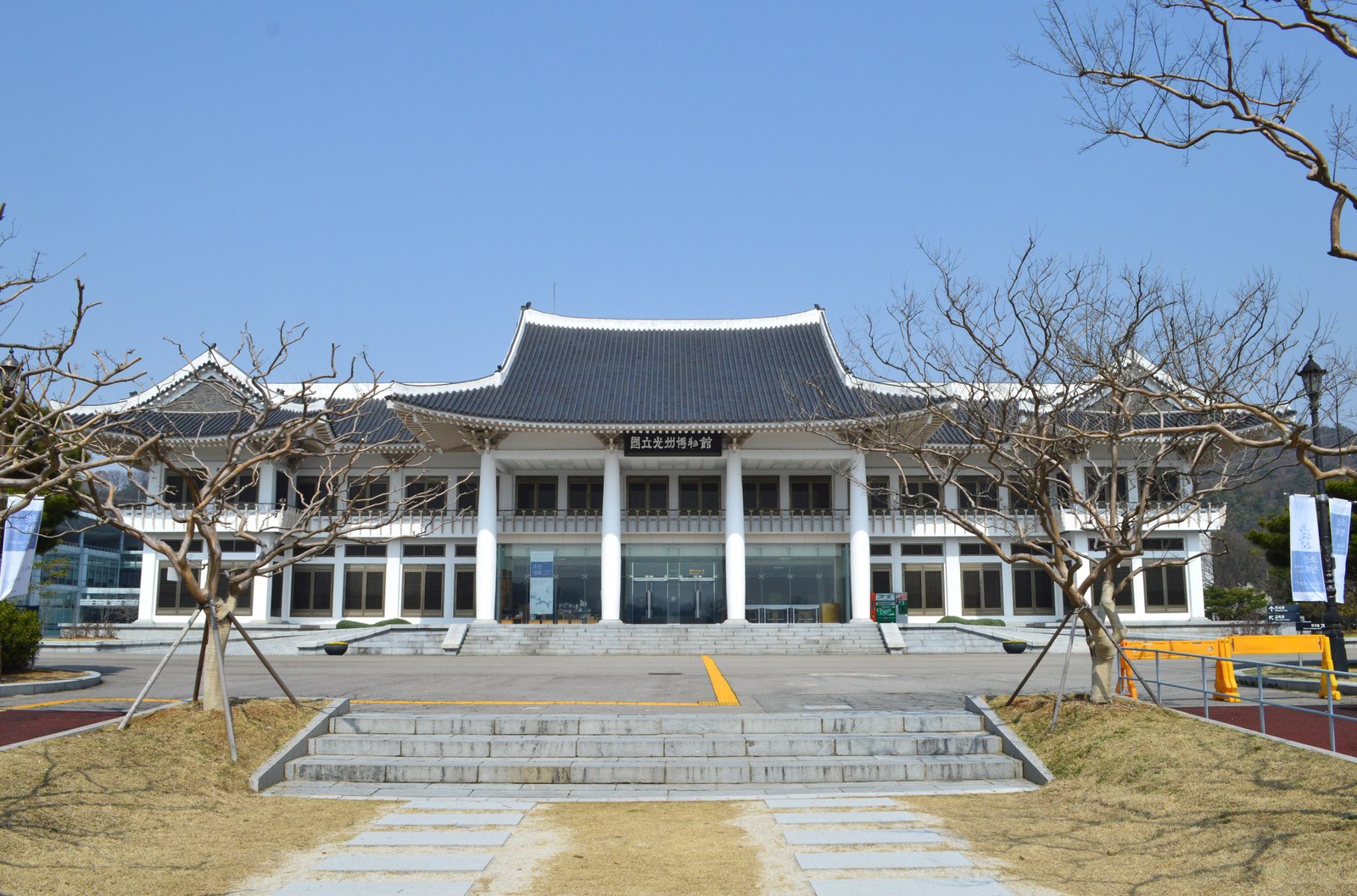 맑은 하늘 아래 흰 기둥이 줄지어 선 박물관 건물 정면이 보인다. 가운데 큰 기와지붕 아래 현판이 있고, 앞에는 넓은 돌계단과 광장, 받침대로 받쳐 둔 잎 없는 나무가 있다.