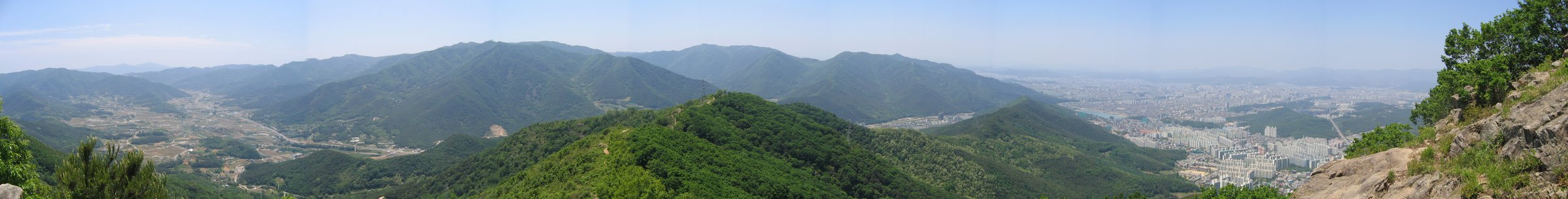 산꼭대기에서 찍은 아주 긴 풍경 사진. 왼쪽은 산줄기 사이 골짜기에 논밭과 마을이 있고, 가운데는 초록 산이 겹겹이 이어지며, 오른쪽으로 가면 아파트와 건물이 빽빽한 큰 도시가 넓게 펼쳐진다.