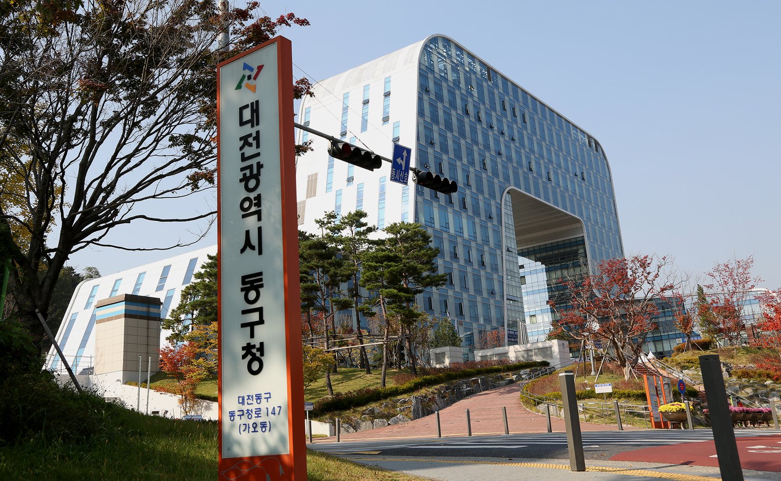 가운데가 커다란 문처럼 네모나게 뚫린 유리 건물 앞에 '대전광역시 동구청'이라고 쓴 주황 테두리 세로 표지판이 서 있다. 둘레에 소나무와 붉게 물든 나무가 있고, 길 위에 신호등이 달려 있다.