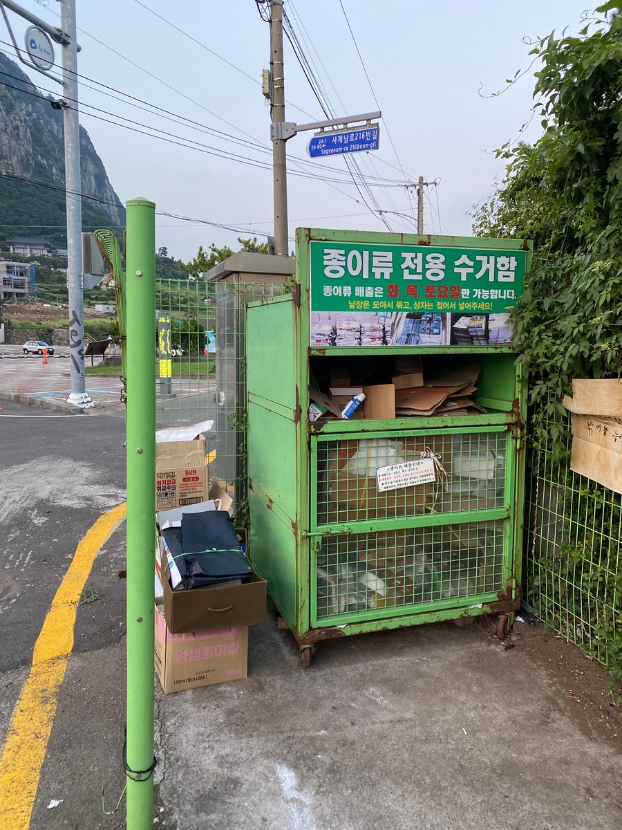 초록색 철제 수거함 위에 종이류 전용 수거함, 종이류 배출은 화, 목, 토요일만 가능합니다라고 쓴 판이 있다. 옆에 종이 상자가 쌓여 있고, 뒤로 큰 바위산이 보인다.