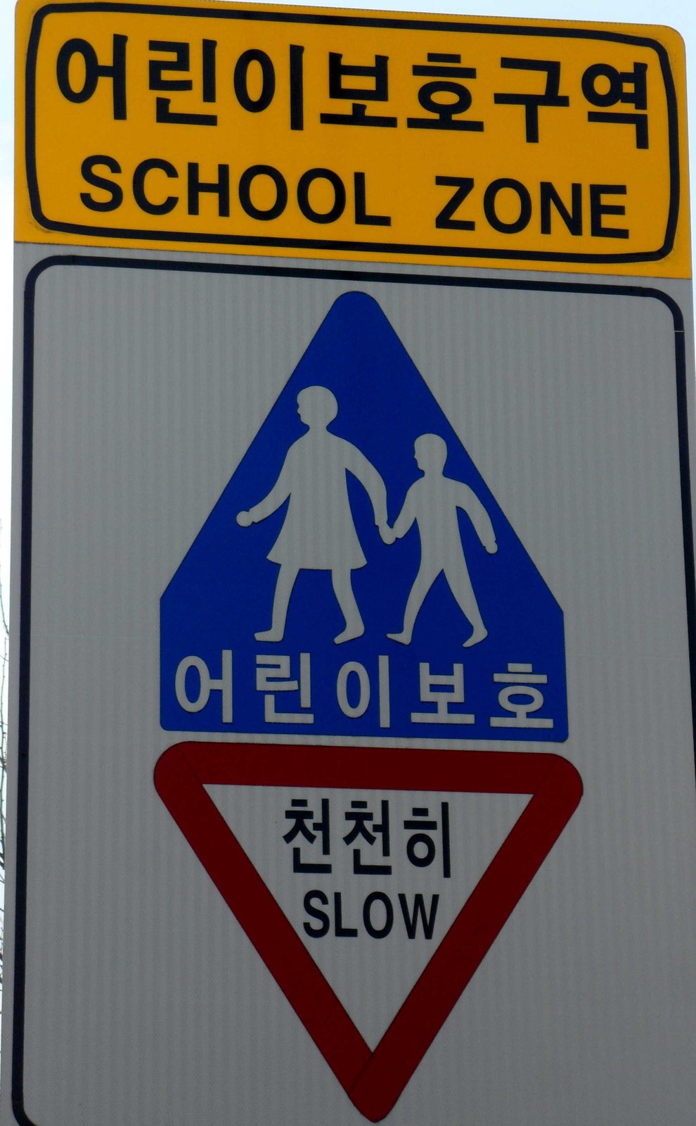노란 판에 '어린이보호구역'이 크게 쓰여 있고 그 아래에 영어 글씨가 있다. 위가 뾰족한 파란 판에는 손을 잡고 걷는 어른과 아이 그림과 '어린이보호' 글씨가 있다. 맨 아래 빨간 테두리의 거꾸로 선 세모 안에 '천천히'가 쓰여 있다.