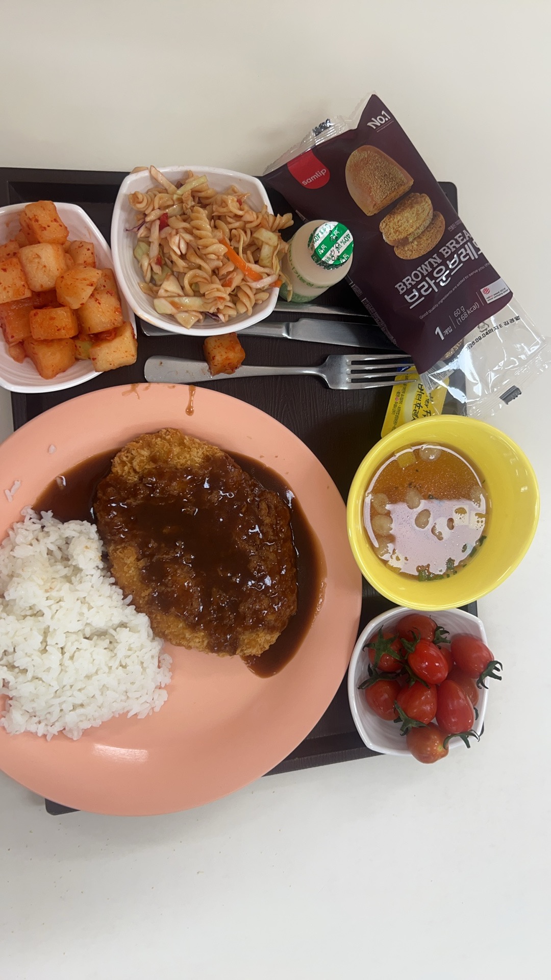 식판 위에 밥과 소스를 얹은 돈가스, 깍두기, 파스타 샐러드, 노란 그릇의 국, 방울토마토, 포장된 빵과 음료가 놓인 급식 한 끼