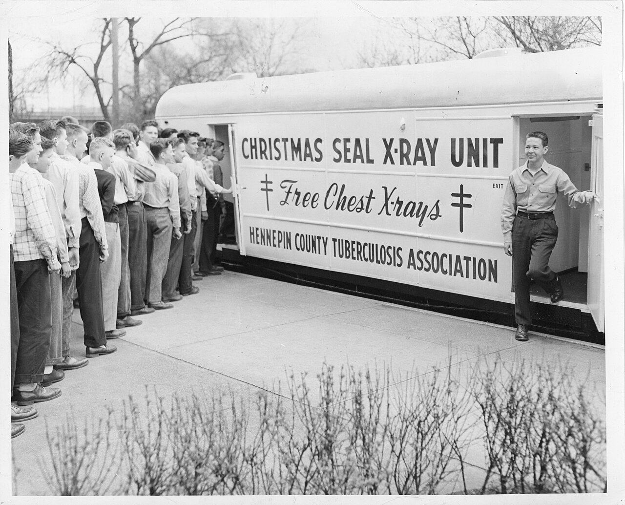 'CHRISTMAS SEAL X-RAY UNIT · Free Chest X-rays · HENNEPIN COUNTY TUBERCULOSIS ASSOCIATION'이라고 적힌 검진 차량 옆에 젊은 남자들이 길게 줄을 서 있고, 촬영을 마친 한 사람이 오른쪽 끝 출구 문으로 내려오는 흑백 사진
